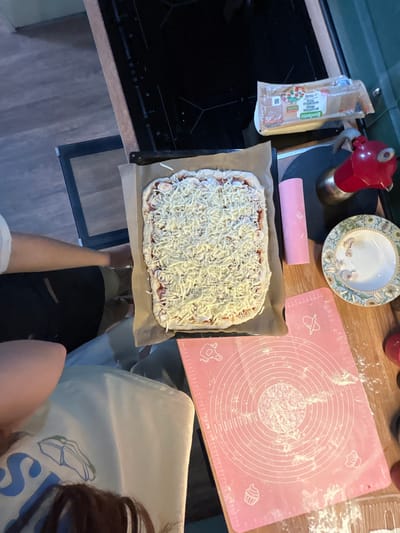 The image depicts a kitchen scene, likely in Shepelyovo, Russia, where a rectangular pizza is being prepared. The pizza, topped with a generous amount of shredded cheese, rests on a baking sheet lined with parchment paper. In the background, a stovetop is visible, and to the right, a red moka pot and a decorative plate with a floral pattern are present. A pink silicone baking mat, dusted with flour and featuring circular guides and icons of kitchen utensils, is prominently placed in the foreground. A person's arm and shoulder are visible on the left side of the frame, suggesting their involvement in the pizza-making process. Another person's head and hair are visible at the bottom of the image, looking down at the baking mat. The overall environment appears to be a home kitchen with wooden flooring and countertops.