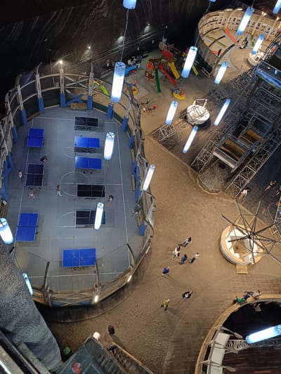 This is an aerial, wide-angle view of the interior of the Salina Turda, a salt mine amusement park located in Turda, Romania. The scene depicts a large, cavernous space with a rough, dark, and textured ceiling. Artificial lighting, in the form of long, rectangular blue tubes, illuminates the area.
In the foreground, a large, enclosed rectangular court with a grey surface is visible. It is marked with white lines and contains multiple blue and black ping pong tables. Several individuals are engaged in playing ping pong, some actively hitting the ball while others stand near the tables. A net encloses this playing area.
Adjacent to the ping pong court, a sandy area features a playground with slides, a merry-go-round, and swings. A few people, including children, are interacting with the playground equipment.
Further into the space, there are elevated walkways, metal scaffolding, and viewing platforms. A circular structure with tiered seating and some exhibits, possibly historical photographs, is also present. Other areas of the cavern show people walking around or sitting, enjoying the recreational facilities. The overall environment is a subterranean complex adapted for leisure activities.