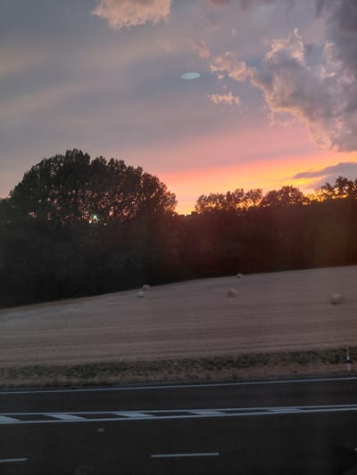 This image captures a rural landscape at sunset, possibly in the vicinity of Ecublens, Switzerland, given the context. The sky displays a gradient of soft pinks and oranges, transitioning into a muted purple, with scattered clouds tinged by the fading sunlight. In the foreground, a dark asphalt road with white lane markings is visible. Beyond the road, a harvested field stretches out, dotted with several round hay bales. A dense line of trees forms a dark silhouette against the vibrant sky, with a notable cluster of trees in the middle ground, partially obscuring the setting sun which emits a bright light through its branches. The overall scene evokes a sense of twilight tranquility.