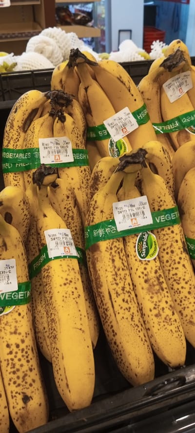 The image shows bunches of overripe bananas with brown spots, secured with green bands labeled "VEGETABLES" and "FRESH" in Japanese. Price tags are visible, some reading "SNPD PIG CRHVG" and "SNPPO PSG CRAVG," with the "Sunpride Cavendish" logo on some. The bananas are arranged in a black plastic crate, suggesting they are on display at a supermarket. Other produce wrapped in white net can be seen in the background.