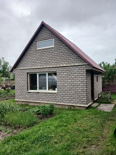 A small, single-story house with a gable roof covered in brown shingles is situated in a green, grassy yard. The house itself is clad in gray brick tiles. There is a large window on the front of the house with white trim and multiple panes, and a smaller rectangular window near the peak of the roof. A wooden door is visible on the side of the house. The surrounding environment includes a patch of garden with green plants and some trees in the background. The sky is overcast and gray. The scene appears to be in a residential area, possibly a dacha or country house, in the town of Sharkawshchyna, Belarus.