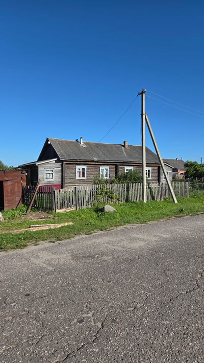 This is a full-frame, eye-level, outdoor, daytime, vertical shot of a rural scene. A long, weathered, wooden house with a gray, corrugated roof is the main subject, viewed from its side. The house has several windows with blue trim. A wooden picket fence runs in front of the house, with some greenery and a large rock visible behind it. A concrete utility pole with wires stands to the right of the house, casting a shadow on the fence. A portion of a cracked asphalt road is in the foreground. The sky is clear and bright blue. The scene appears to be in a rural area of Zarubino, Russia.