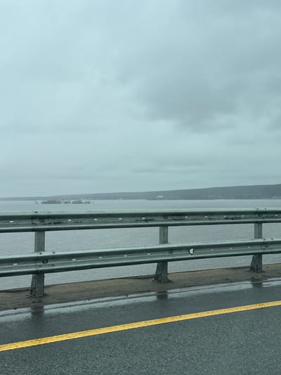The image shows a view from a road on a foggy day. To the right of the frame is a grey asphalt road with a yellow line marking the edge. A metal guardrail runs along the edge of the road, separating it from the body of water. The body of water appears to be a lake or river, with a shoreline visible in the distance. The sky is overcast and grey. The location is presumed to be near a body of water in an urban or suburban area, based on the roadway and guardrail and a distant treeline and possibly buildings. There is no clear evidence to confirm that this image is located in Grebeni, Russia.