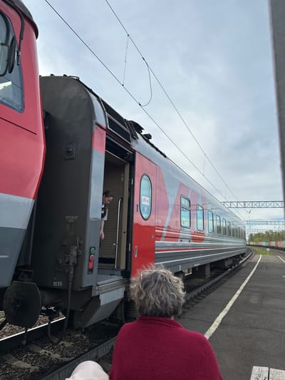 A red and grey train stands on tracks at a station in Кечёво, Russia. The train's door is open, revealing a woman inside the train car. A person with grey hair and wearing a red sweater is standing next to the train, looking toward the train and the woman inside. The sky is overcast, and the station platform is visible next to the tracks. Overhead power lines are also visible.
