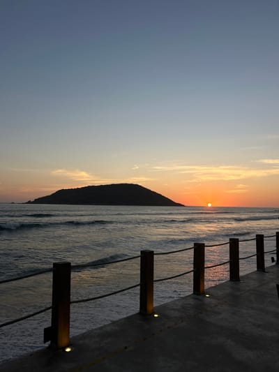 This image captures a serene sunset over the Pacific Ocean, likely in Mazatlán, Mexico, as viewed from an elevated boardwalk or pier. The sun, a bright orange orb, is positioned low on the horizon, casting warm light across the scene. A prominent, dark silhouette of a mountain or large hill stands against the vibrant sky, partially obscuring the setting sun and adding depth to the landscape.
The sky transitions from a deep, cool blue at the top to hues of soft orange and yellow near the horizon, with delicate, wispy clouds illuminated by the setting sun. The ocean stretches out, its surface characterized by gentle waves that reflect the warm, golden light of the sunset, creating shimmering patterns on the water.
In the foreground, a dark, textured concrete structure, possibly a walkway or viewing platform, runs along the right side of the image. It features a railing composed of several sturdy wooden posts with ropes or thick cables strung between them. Each wooden post has a small, circular light fixture embedded in its side, casting a subtle, warm glow onto the concrete and adding an inviting element to the structure. No people are visible, emphasizing the tranquility and natural beauty of the moment. The overall impression is one of peaceful contemplation at the close of day. There is no visible text in the image.
