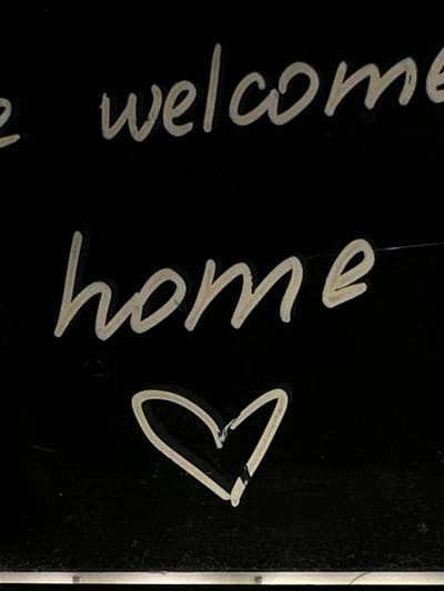 The image shows a close-up, dimly lit view of white handwritten text on a black background. The text is stylized and cursive, with the words "e welcome" appearing on the top line and "home" on the second line. Below the text, a white heart shape is drawn. The handwriting appears to be chalk or a similar medium. The background shows some texture and slight imperfections, suggesting a chalkboard or a similar dark surface. The lighting is focused on the writing, creating a somewhat dramatic effect. There are no people or specific city landmarks visible. The location in Podilsk, Ukraine, is not discernible from the image itself.
