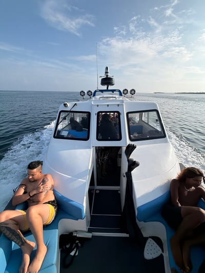 The image shows two shirtless young men relaxing on the deck of a motorboat. One man is lying on his back on a blue seat, wearing yellow swim trunks and displaying tattoos on his arms and legs. The other man is sitting with legs extended on a blue seat in the background, with his back to the camera. The boat has a white exterior, with an enclosed cabin, and is moving through the water, as evidenced by the wake. The sky is partly cloudy. Based on the mention of Dhigurah, Maldives, the location is likely in the ocean near the island.