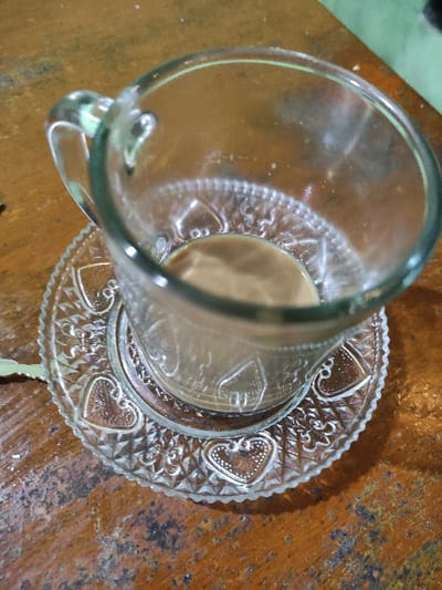 This image captures a close-up, high-angle view of a clear glass mug containing a residual amount of light brown liquid. The mug, which has a C-shaped handle on its left side and an embossed pattern on its interior base, is centered on a matching clear glass saucer. The saucer features a scalloped edge and an intricate raised pattern of hearts and geometric forms. Both items rest directly on a flat, horizontal wooden surface characterized by varied reddish-brown to dark brown tones, visible grain, and surface wear. A blurred green element is partially visible in the upper right background. No individuals or active interactions are present in the frame. The location depicted is Staufenberg, Germany.