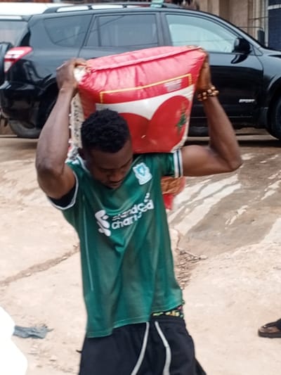 The image depicts an outdoor scene in Awada, Nigeria. A young adult male with short, dark curly hair, wearing a green short-sleeved Liverpool FC football jersey sponsored by "Standard Chartered" and black shorts with white side stripes, is shown carrying a large, red and white sack on his head. His arms are raised to steady the sack, which features a prominent red heart-like graphic on a white background and a green plant emblem. A multi-beaded bracelet is visible on his right wrist. His gaze is directed downwards. In the background, a black Toyota RAV4 SUV is partially visible. The ground is an uneven, light-colored surface, appearing to be concrete or compacted dirt. A dark piece of fabric rests on the ground to the lower left, and a human foot in a brown sandal is partially visible to the lower right.