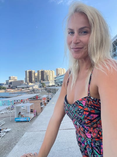 A woman with blonde hair and blue eyes is taking a selfie. She is wearing a colorful patterned swimsuit. Her face is close to the camera, and she is smiling. In the background, there is a clear blue sky with a crescent moon visible. Below the sky, there is a city skyline with tall buildings. There is also a beach or resort area with lounge chairs, umbrellas, and people relaxing. A sign with Cyrillic text that reads "АФРОКОСЫ" is visible on a small building. The lighting suggests it is daytime, likely late afternoon or early evening given the presence of the moon. The scene appears to be a vacation or leisure setting, possibly a rooftop terrace overlooking a beach resort.