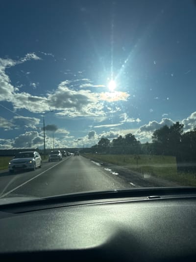 The image shows a view from the inside of a car, looking out at a road with traffic. The sun is shining brightly in a blue sky with scattered clouds. The road appears to be a highway with multiple lanes and cars driving in the same direction. There are trees and green fields on either side of the road. A traffic sign is visible on the right side of the road. The car's dashboard is visible in the foreground. The location is identified as Polivanovo, Russia.