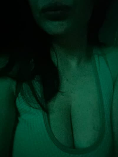 The image is a close-up, low-light, green-tinted photograph of a person's upper body. The subject is wearing a white garment with a textured pattern, possibly a tank top or a bra, and the décolletage area is visible. Dark hair frames the left side of the image. The lighting is dim, with shadows obscuring some details of the face and upper body. The scene appears to be indoors, with no specific city or location identifiable within the frame itself.