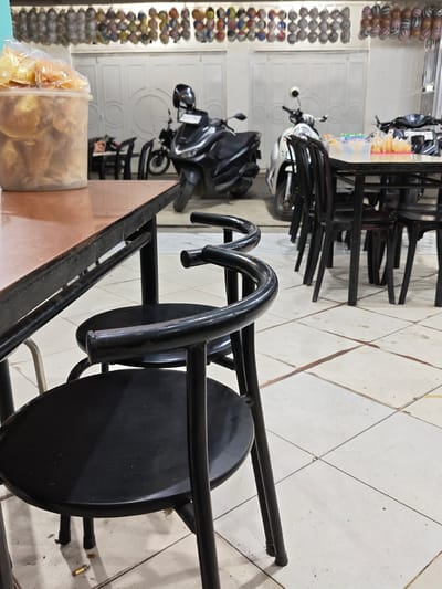 This is a medium-shot, eye-level, interior photograph of a simple cafe or eatery, likely located in Blangmetimu, Indonesia. The scene is dimly lit, with the primary light source appearing to be from above and behind the viewer.

In the foreground, a round, black plastic chair with a curved metal backrest is prominently featured, positioned at an angle. Its metal frame shows signs of wear, with some scuff marks visible. Another similar chair is partially visible behind it. To the left, a portion of a rectangular table with a reddish-brown laminate top and black metal legs occupies the left side of the frame. A clear plastic container filled with what appear to be light-colored, fried crackers or snacks sits on the table, partially obscuring the view beyond.

In the mid-ground, the main seating area of the establishment is visible. Several identical black metal chairs with round seats are arranged around tables that are mostly obscured. Behind these tables, two motorcycles are parked. The one on the left is a black scooter, and the one on the right is a silver and black scooter. Both have helmets resting on their seats.

The background features white double doors with decorative circular patterns. Above these doors, a row of what appear to be colorful, netted bags containing small objects, possibly snacks or trinkets, are hung along a wall. The floor is tiled with square, off-white tiles, some of which appear to have a reddish-brown stain. Dust and debris are visible on the floor and on some of the furniture. The overall impression is that of a casual, functional dining space.