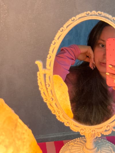 The image shows a young woman looking into an ornate, oval mirror. The mirror is framed in gold and has decorative scrollwork. Her reflection is visible in the mirror, showing her face tilted, with her hand resting on her cheek. She is wearing a pink long-sleeved shirt. She is holding a pink phone, which is partially obscuring her face. Her nails are painted with a glittery polish. The background in the reflection is a blue wall. The surrounding environment appears to be indoors, with a textured gray wall to the left of the mirror and a yellow, textured object to the bottom right. The lighting suggests it might be evening or indoors with artificial light. There is no visible text in the image. The setting is likely a personal space, such as a bedroom or bathroom, in Tliustenkhabl', Russia.