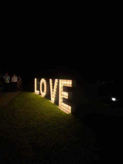 The image captures a nighttime scene in Baliovtsi, Bulgaria, where a large, illuminated sign spelling out "LOVE" stands prominently on a grassy area. The sign, composed of block letters with exposed bulbs, casts a warm glow on the surrounding grass. To the left of the sign, a small group of people are gathered. One individual, dressed in a light-colored shirt, stands with their back to the camera. Beside them, another person in a similar shirt is also standing. Further to the left, a woman with a patterned dress is seated on a stool. The background is dark, indicating the absence of ambient light, with faint lights visible in the distance, suggesting the presence of buildings or other structures. The overall atmosphere is serene and festive, likely part of an outdoor event.