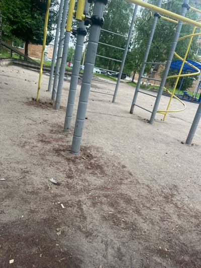 The image depicts an outdoor sports playground in Usogorsk, Russia, with a focus on the sandy ground and supporting structures of the equipment. A row of grey metal poles, part of a climbing frame, dominates the foreground. Some of these poles have yellow accents. The ground is sandy and uneven, littered with small brown debris, possibly pine cones or wood chips, and a single white feather near the bottom left. In the background, lush green trees form a dense canopy. Beyond the trees, residential buildings are visible, typical of a Russian urban environment. The overall impression is of a functional but somewhat unkempt public space.