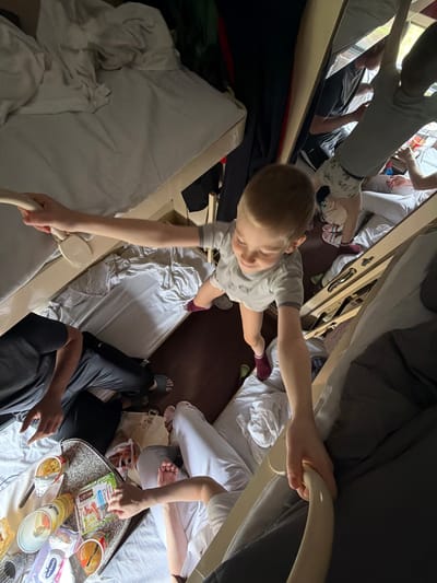 This high-angle shot captures a busy, communal interior space, strongly suggesting a train cabin or a similar bunk-bed arrangement, given the cramped quarters and stacked sleeping areas. The scene appears to be taking place during the daytime, illuminated by natural light from an unseen source.

In the center, a young boy with light brown hair, wearing a light-colored t-shirt and dark socks, is actively moving. He is reaching with both hands, holding onto parts of the bunk bed structure, either climbing down or navigating the space between berths. His expression seems focused.

Around the boy, several other individuals are visible, though mostly partially. Above him, a person is seen in grey pants and a light top, their legs extending. Below and to the right, a person in dark pants and a t-shirt is seated, with another individual in white pants and a white top nearby. They appear to be engaged in eating or snacking.

On a small, irregularly shaped tray placed on a bed to the right, various food items are arranged. Visible packaging includes a green pouch labeled "ИНДЮШИНЫЕ СОСИСКИ МОЛОЧНЫЕ" (Turkey Milky Sausages) with the number "5", indicating a food product. A yellow cup with "РОЛЛТОН" (Rollton), a brand of instant noodles, is also present, along with a white container possibly for cream cheese or a similar dairy product, with text that looks like "Watsushi". Other items include what appear to be yogurts, bread, and various snacks.

The beds are covered with white and grey bedding, some of it rumpled. Personal items like socks and slippers are scattered on the floor or near the beds. The setting evokes a sense of travel and shared space, consistent with a long-distance journey, possibly through Russia, given the Bobrov context. The overall atmosphere is lively and informal, centered around travel and shared meals.