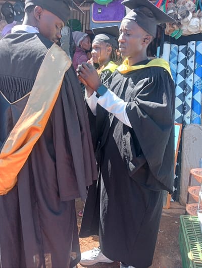 The image depicts three individuals in the city of Waseges ward, Kenya, wearing black graduation gowns and mortarboard caps in an outdoor setting. In the foreground, two males stand on reddish-brown ground. The male on the left has his back partially to the camera, wearing a black gown with an orange/gold stole. The male on the right, facing left, wears a black gown with a yellow stole over a white long-sleeve shirt, a smartwatch on his left wrist, and has his hands clasped together at chest level. A female, also in a black gown, mortarboard cap, and yellow stole, is positioned in the mid-ground, looking towards the left. A fourth individual, wearing a grey beanie, is partially visible in the background. The background features a market-like environment with various items displayed: hanging hats/caps on the left, and rolls of patterned fabric (blue, white, black geometric designs) and a brown multi-tiered shelf on the right.