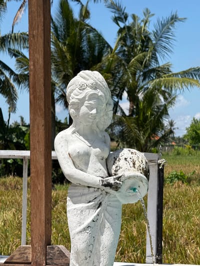 A sideways, eye-level shot shows a white, concrete statue of a woman holding a water vessel that emits two streams of water, located in Gubug, Indonesia. The statue rests on and is surrounded by a wooden and metal support structure. The backdrop features palm trees and tall grass under a partially cloudy sky.