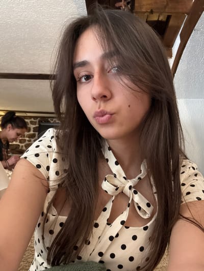 A young woman with long, dark brown hair and fair skin is taking a selfie. She is wearing a white dress with black polka dots and a bow at the neckline. Her lips are pursed, and her gaze is directed towards the camera. In the background, another woman with her hair pulled back in a bun is visible, facing away from the camera and looking down at something. The environment appears to be indoors, with a textured ceiling and stone walls. The setting is implied to be in the city of Smilyan, Bulgaria.