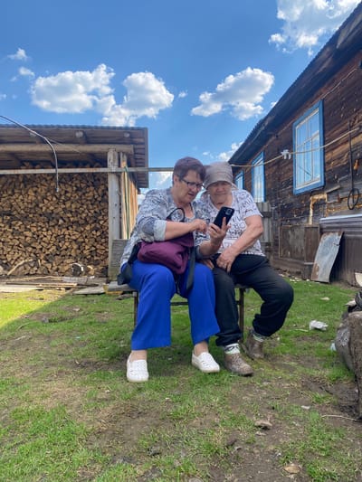 Two women are seated on a bench outdoors. One woman, wearing a white patterned shirt and a headscarf, is holding and looking at a smartphone. The other woman, wearing a light patterned blouse, glasses, and blue pants, is looking at the phone. They are sitting in front of a woodpile and an old wooden structure with a blue-framed window. The location appears to be a rural setting, with grass and muddy ground. The sky is visible, with blue sky and white clouds. Based on the information provided the location is Узяр, Russia.