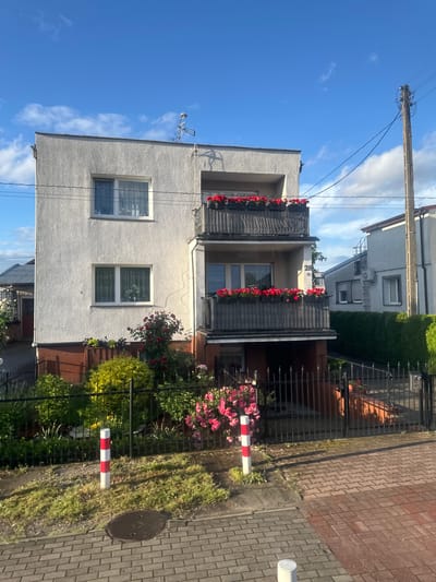 This image shows a two-story building in Krosno Odrzańskie, Poland. The building is white with a red brick foundation. It has two balconies with red flowers in planters. The property is enclosed by a black metal fence. In the foreground, there is a sidewalk made of brick pavers, a manhole cover, and a red and white striped pole. The sky is blue with some white clouds. The scene appears to be set in a residential area.