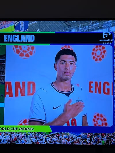The image shows a television screen displaying a promotional image for the "WORLD CUP 2026". A male athlete, wearing a white jersey with a Nike logo and the number 10, is prominently featured. He is placing his hand over his chest. The background is light blue with several red and white patterned circular motifs, resembling roses or stylized footballs. The words "ENGLAND" are displayed in large blue letters on a black bar at the top of the screen, and "AND" and "ENG" are visible in large red letters on the blue background. In the top right corner, there is a logo for "antenaPLAY LIVE". Below the athlete, a crowd of spectators is visible. The image is framed by a television bezel. The location in Florești, Romania is not discernible from the provided image.