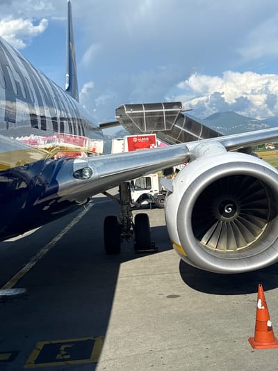 The image shows a close-up view of the engine and wing of an airplane on the tarmac at an airport. The airplane is a large commercial jet with a distinctive blue and silver livery. The engine is a turbofan, with visible fan blades inside the cowling. A jet bridge is connected to the aircraft, and a ground vehicle, possibly a baggage tug, is parked nearby. In the background, mountains are visible under a cloudy sky. The scene appears to be set during the daytime, with strong sunlight casting shadows. The ground is asphalt, marked with yellow lines and the letter "E" painted in a dark blue rectangle. An orange traffic cone is visible in the foreground. The location is identified as Kruja, Albania.