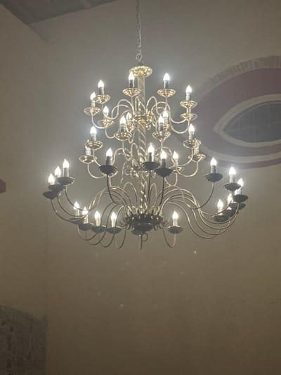 A chandelier with multiple tiers of lights hangs from a ceiling. The fixture features ornate metalwork with curling arms supporting small, bright bulbs. In the background, there's a decorative oval shape outlined in shades of gray. The room has a warm, off-white tone. A glimpse of what appears to be a white, lacy material is visible in the lower right corner.