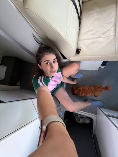 The image depicts a woman and a small dog in what appears to be the interior of a vehicle or private aircraft. The woman is seated on the floor, her legs bent and extended. She is wearing a green and pink t-shirt and blue socks. A small, reddish-brown dog, possibly a poodle mix, is also on the floor, near the woman's legs. In the background, there are cream-colored leather seats with black seatbelts, suggesting a luxurious interior. The floor is a dark, speckled material. There are also some storage compartments visible. The scene is captured from a high-angle perspective, as if the photographer is holding the camera above their head. The location in Nea Moudania, Greece is not discernible from the image itself.