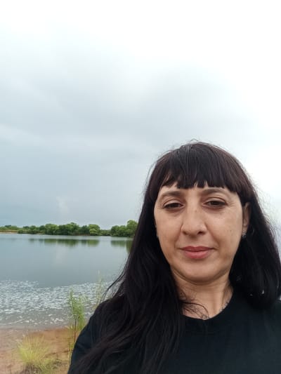 A woman with dark hair and bangs, wearing a black t-shirt, takes a selfie outdoors with a lake and trees in the background under an overcast sky. The woman's face is turned slightly towards the camera, and she has on light lipstick and earrings. The water in the lake is calm, reflecting the trees on the opposite bank. The ground in the foreground appears to be sandy and dry, with some sparse vegetation. The location is Vasil'evka, Russia.