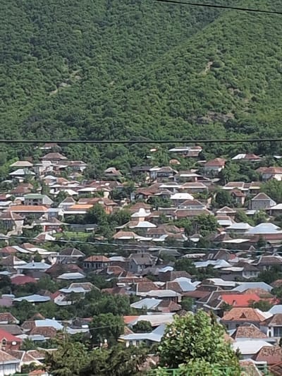 This media file captures an aerial view of a densely populated area nestled against a backdrop of lush green mountains. The scene is dominated by a multitude of buildings, characterized by their distinct roof designs, many of which are covered in red or brown tiles, interspersed with white and lighter colored facades. The architecture suggests a historic or traditional settlement, with buildings appearing to be tightly clustered together. Green foliage, likely trees and vegetation, is abundant, not only in the surrounding mountains but also interspersed among the buildings, indicating a harmonious integration of nature and human settlement. The lighting and shadows suggest it is daytime, likely late morning or afternoon, with the sun casting distinct shadows on the roofs and landscape, though no specific weather conditions are discernible. The perspective is from a high vantage point, possibly a drone or a helicopter, offering a sweeping panorama of the town and its natural surroundings. There is no visible text in the image. The overall impression is one of a vibrant community embedded within a scenic, mountainous landscape, possibly in a location like Şəki, Azerbaijan, known for its historical architecture and natural beauty.