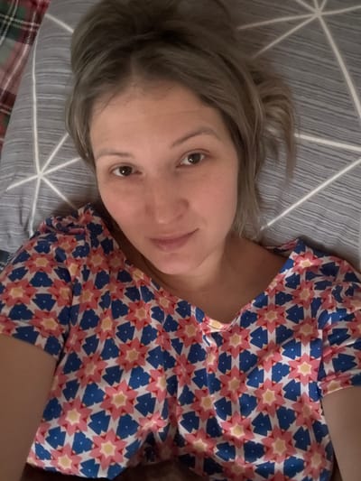 A woman with blonde hair lies on a patterned grey pillow. She wears a brightly colored shirt with a geometric design of blue, red, and white. She gazes into the camera with brown eyes. Her complexion appears fair. The bedding includes a grey striped pattern near her head and a glimpse of plaid on the bottom left of the frame.