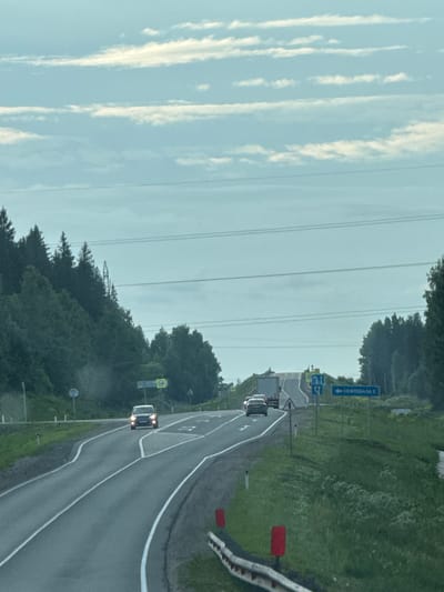 The image depicts a two-lane asphalt road curving through a forested landscape under a cloudy sky. A white SUV is approaching from the opposite direction, its headlights illuminated. A truck is visible further ahead on the same road, also traveling in the opposite direction. On the right side of the road, a blue directional sign points to the left, indicating "НЕФТЕБАЗА 2" (NefteBaza 2). The surrounding environment is characterized by dense green trees on both sides of the road, with a grassy embankment on the right. Power lines are strung across the upper portion of the frame. The scene suggests a rural or semi-rural setting.