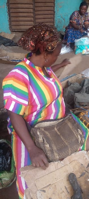 This image depicts an informal market scene in Ilogbo Eremi, Nigeria, featuring two women. In the foreground, a woman of medium-dark complexion is seated, oriented away from the viewer, with her head turned to her right, observing items on the ground. She wears a short-sleeved top with vertical rainbow stripes (red, orange, yellow, green, blue, purple, white) and a brown headwrap adorned with metallic patterns. Small silver hoop earrings are visible. A brown, patterned sling bag rests across her torso. Her right hand is placed on a stack of worn, discolored cardboard pieces lying on the ground. To her left, a green woven sack contains dark, possibly bagged items. On the cardboard, a single dark, elongated object, possibly a dried fish, is present. In the background, a second woman with a darker complexion is seated on the ground, wearing a patterned blue and brown dress. She is actively handling white granular material, likely rice or garri, from several clear plastic bags. A blue and white patterned cloth is partially visible beneath her. The immediate environment includes a weathered wall painted a light blue or teal, exhibiting significant paint loss to reveal reddish-brown substrate, and a window fitted with horizontal wooden slat shutters. The ground appears earthy and uneven.