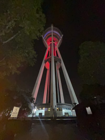 The image shows the Alor Setar Tower at night in Alor Setar, Malaysia. The tower is illuminated with red and blue lights. Trees are visible in the foreground and on the sides of the image. A circular building is at the base of the tower. A sign with text is visible on the left side of the image. The scene is dark, indicating it is nighttime.