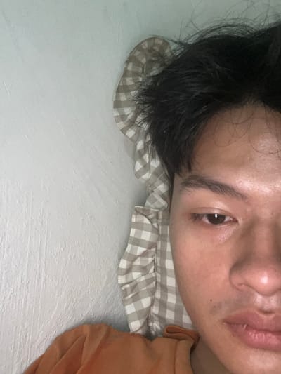 A close-up, overhead shot captures a person's face, partially obscured by dark, messy hair, resting on a light-colored, checkered pillow. The person is looking towards the camera, their right eye clearly visible. The background consists of a textured, light-colored wall. A portion of an orange garment is visible in the lower left corner. The scene appears to be indoors, possibly in a bedroom. The specific location in Serdang Kulon, Indonesia, is not discernible from the image itself.