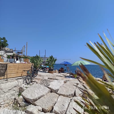 The image depicts a sunny day at a rocky beach with a view of the sea. In the foreground, large, light-colored rocks form the uneven terrain. To the left, a wooden structure with a slatted front is visible, and a bicycle is parked against it. Further back, a second bicycle is also visible. Several large potted cactus plants are strategically placed. In the middle ground, several people are relaxing on the beach, some under umbrellas, while others are sitting or lying on towels. A blue and a green umbrella provide shade. The sea is a deep blue, with gentle waves. In the background, there are some structures that appear to be part of a seaside establishment, including what looks like a fishing platform or pier with a net. The sky is a clear, bright blue. The date stamp "22.06.2026 10:41" is visible in the bottom right corner. Based on the context and the presence of a structure resembling a "trabucco," this scene is likely located in Trani, Italy, a coastal city known for such traditional fishing platforms.
