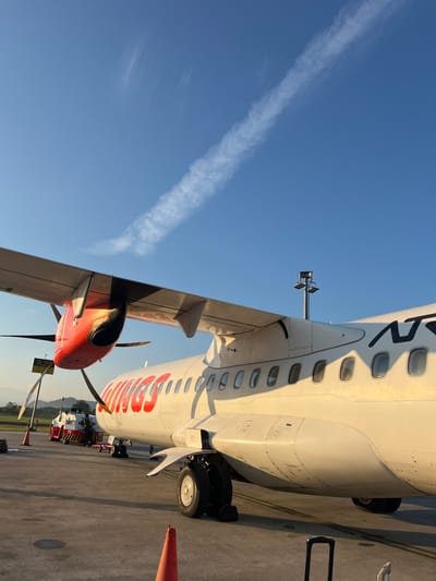 The image shows a partially visible aircraft on an airport tarmac. The aircraft, predominantly white with red accents and lettering, features a turboprop engine with a red nose and black propellers. The sky is clear blue, marked with a white contrail. Ground support vehicles, including a baggage tug, are visible near the aircraft. The photo was taken on an airport. No specific location within Baji Mangngai, Indonesia, can be determined based solely on the information contained in this image. There are no people visible in the image.