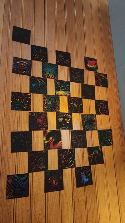 The image shows a collection of scratch art pieces arranged on a light brown wood-paneled wall. The wall is likely an interior wall, given the lack of exterior features. The scratch art pieces are small, square or rectangular, and feature various colorful designs revealed by scratching away the black surface. The designs include abstract patterns, floral motifs, faces, and other recognizable shapes. The composition of the arrangement is somewhat grid-like but not perfectly aligned. There is no indication that the image was taken in Jackson, United States of America. There are no people in the image.
