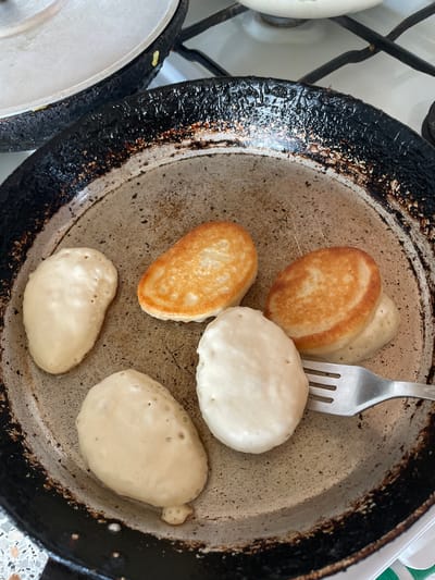 The image depicts a frying pan on a stovetop, likely indoors. The pan contains several pancakes in various stages of cooking, some golden brown and others pale. A metal fork is present in the pan, seemingly being used to maneuver the pancakes. The pan appears well-used with a blackened exterior. The setting is likely a residential kitchen. The city of Talmenka, Russia is only a location detail provided by the user and is not visible in the image.