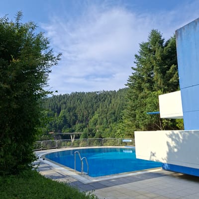 The image shows an outdoor swimming pool with bright blue water, surrounded by a tiled deck. A metal fence with railings borders the pool area. Beyond the pool, a dense, green forest covers a hillside. The sky is blue with scattered white clouds. The sun appears to be casting shadows, suggesting it is daytime. The scene is set in a natural environment, likely a resort or recreational area. There is a sign on the fence with some text, but it is not clearly legible. The overall impression is one of a serene and inviting outdoor setting.