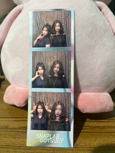 A photostrip featuring three pairs of poses by two young women is presented. The photostrip is placed on a wooden surface, with a large, light pink plush toy to the left and behind it. The background of each photo frame consists of a brown, textured curtain.
In the top panel, the woman on the left, facing forward, has her left hand on her cheek, and the woman on the right, facing slightly left, looks towards the camera. Both are wearing dark tops.
The middle panel shows the woman on the left, facing forward with her hands on her cheeks, and the woman on the right, looking towards the camera. Both are wearing dark, off-the-shoulder tops with necklaces.
In the bottom panel, both women are making "peace" signs with their fingers. The woman on the left is facing forward, and the woman on the right is looking towards the camera. They are both wearing dark tops. The photostrip has the text "SNAPLAB ODYSSEY" printed at the bottom in white, stylized lettering. The location of Pasir Gadung, Indonesia, is not discernible from the image.