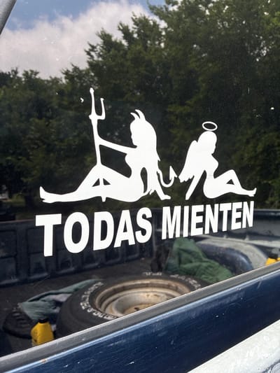 A white decal on a dark surface, likely a vehicle window, depicts two stylized female figures and text. The figure on the left has devil horns, a pitchfork, and a tail, representing a devil. The figure on the right has a halo and wings, representing an angel. Between them, also in white, are the words "TODAS MIENEN". The background consists of green trees and a cloudy sky. The lower part of the image shows a portion of a vehicle bed with a spare tire and a green cloth. The location is Balch Springs, United States of America.