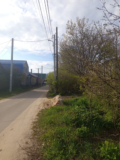 This image depicts a static outdoor scene of a semi-rural or residential area, likely in Svkh Voronezhskiy, Russia. The scene features a narrow, unpaved road or path on the left, next to a wider dirt or sand track on the right.
Along the right side of the track, there's a dense strip of vibrant green grass, bushes, and trees. The trees show signs of early spring, with fresh green leaves emerging and some small white buds or blossoms visible. The vegetation continues into the background, forming a lush green boundary.
On the left side of the road, several utility poles with multiple power lines stretch upwards against a partially cloudy sky. Further along the road, a cluster of houses or buildings can be seen. One prominent house has a distinctive bright blue roof, while another in the distance features a green roof.
The sky is a mix of clear blue patches and scattered white clouds. The lighting suggests it is daytime, with natural light illuminating the scene. No people or specific activities are occurring. There is no visible text in the image.