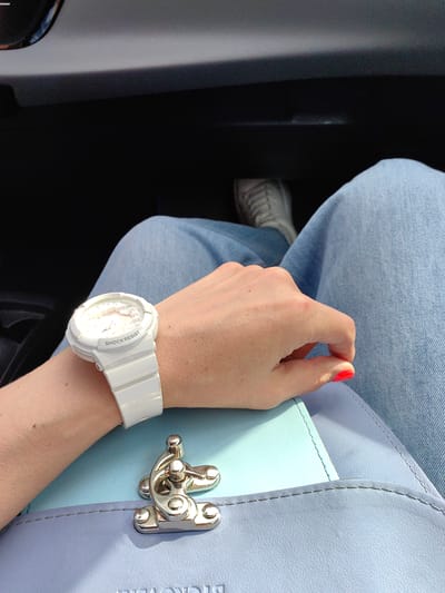 This image depicts a close-up, first-person perspective view of a person seated inside a vehicle, located in Негарэльскі сельскі Савет, Belarus. The subject's left hand, with a light skin tone and visible freckles on the wrist, wears a white Casio Baby-G watch with 'SHOCK RESIST' embossed on the bezel. The thumbnail is painted a vibrant orange/coral, and a small reddish mark is present on the index finger. The hand rests on a two-tone bag, featuring a light blue upper flap and a light purple lower body, secured by a prominent silver-toned metal clasp. The brand name 'BICKOVEM' is embossed on the lower purple section of the bag. The person's lap is covered by light blue denim jeans. In the blurred background, a light-colored sneaker with laces is partially visible on the vehicle's floor, against dark interior panels.