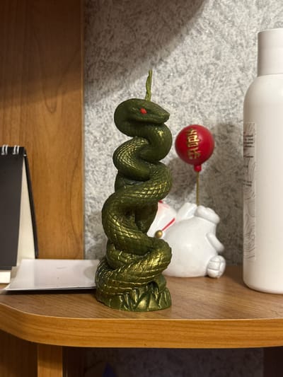 The image shows a snake-shaped candle with a green and gold finish, coiled around itself, with red eyes. The candle is positioned on a wooden shelf. Behind the candle, a white ceramic figure resembling a rabbit is visible, holding a red balloon with gold Chinese characters on it. To the right of the candle is a white bottle with a label. To the left of the candle, a black and white object with a spiral binding is partially visible. The background consists of a textured gray wall and a wooden panel. The scene appears to be an indoor setting, possibly a shelf in a room. The location is identified as Масаловка, Russia.