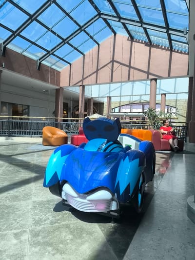 The image depicts the interior of a shopping mall in Montijo, Portugal, likely the Freeport Lisboa Fashion Outlet. In the foreground, a bright blue, cartoonish toy car designed for children is parked on a marble-tiled floor. The car features a cat-like mask on its front and stylized lightning bolt patterns. Behind the toy car, a seating area is visible, with a woman in a red dress sitting on a burgundy sofa, holding a red handbag and looking to the right. The mall features a large, multi-pane glass ceiling that allows natural light to flood the space, casting shadows on the floor. The background reveals more of the mall's architecture, including brown pillars, railings, and shops.