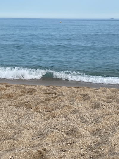 The image depicts a tranquil beach scene with the gentle lapping of waves against the shore. The foreground is dominated by a textured expanse of sand, marked with the subtle imprints of receding water and exhibiting a fine, granular composition. A small wave, characterized by its white foam crest and clear, blue-green water, is captured in the process of breaking onto the beach. The middle ground consists of the calm, blue sea, stretching horizontally across the frame and reflecting the soft, pale sky. A single, small yellow buoy is visible in the distance, floating on the surface of the water, adding a subtle point of color to the vastness of the sea. The horizon line is sharp, dividing the expanse of the ocean from the clear, pale blue sky. The location is identified as the beach of Vilassar de Mar, Spain.
