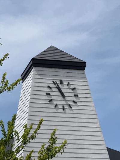 The image presents a striking upward view of a modern clock tower against a clear, partly cloudy blue sky. The tower, located in Fujikawaguchiko, Japan, is constructed with light-colored, vertically ribbed panels, likely white or light gray, which create strong linear patterns and subtle shadows. Its corners and top edges are defined by sleek dark grey or black sections, adding a contemporary contrast to the design.
A minimalist clock face adorns one side of the tower. Instead of numbers, it features bold black rectangular markers for the hours. The clock hands, also black, indicate a time of approximately 3:45, with the minute hand pointing towards the nine o'clock position and the hour hand positioned between the three and four o'clock markers.
The sky above is a vibrant light blue, traversed by delicate, wispy white clouds, suggesting bright daylight. In the foreground, from the lower right and bottom edges of the frame, vibrant green tree branches with abundant leaves partially frame the base of the tower. This low-angle perspective accentuates the tower's height and angular geometry, juxtaposing its modern design with the natural elements of the sky and foliage. There is no visible text in the image.